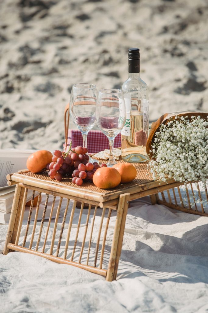 Outdoor picnic setup on a sandy beach with wine, grapes, and flowers.